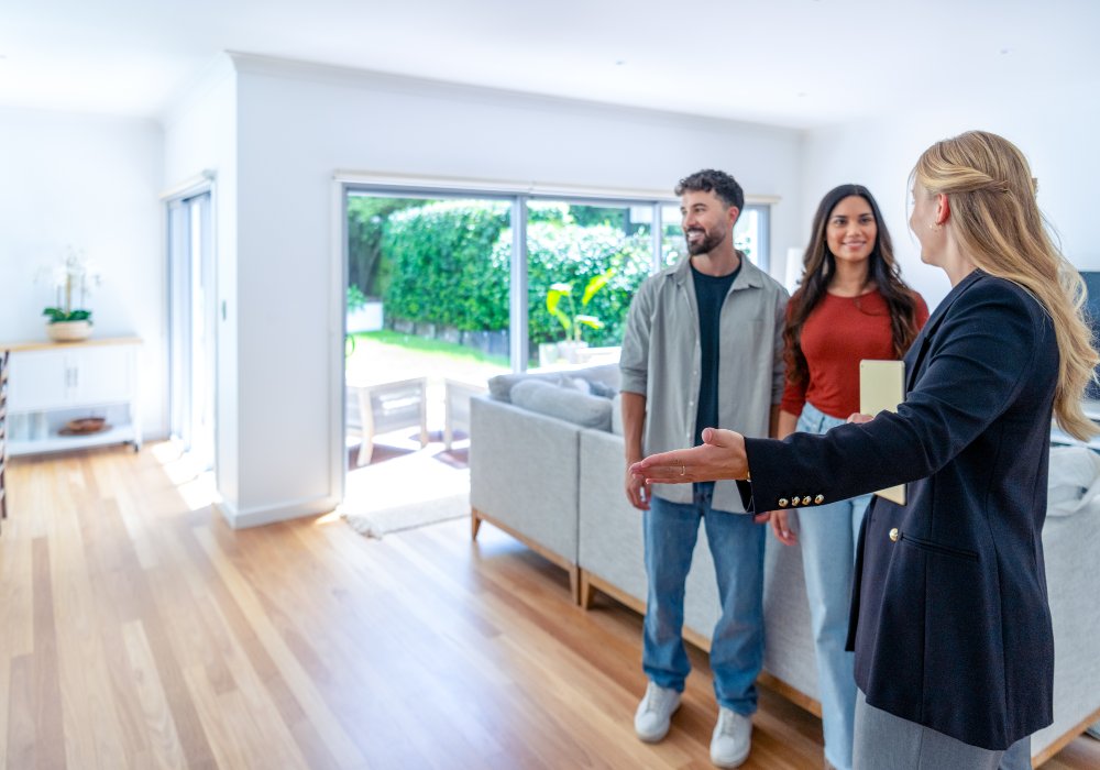 Real Estate agent showing a couple through a new house. They are standing in the living room. Outdoor area and yard can be seen through a window.